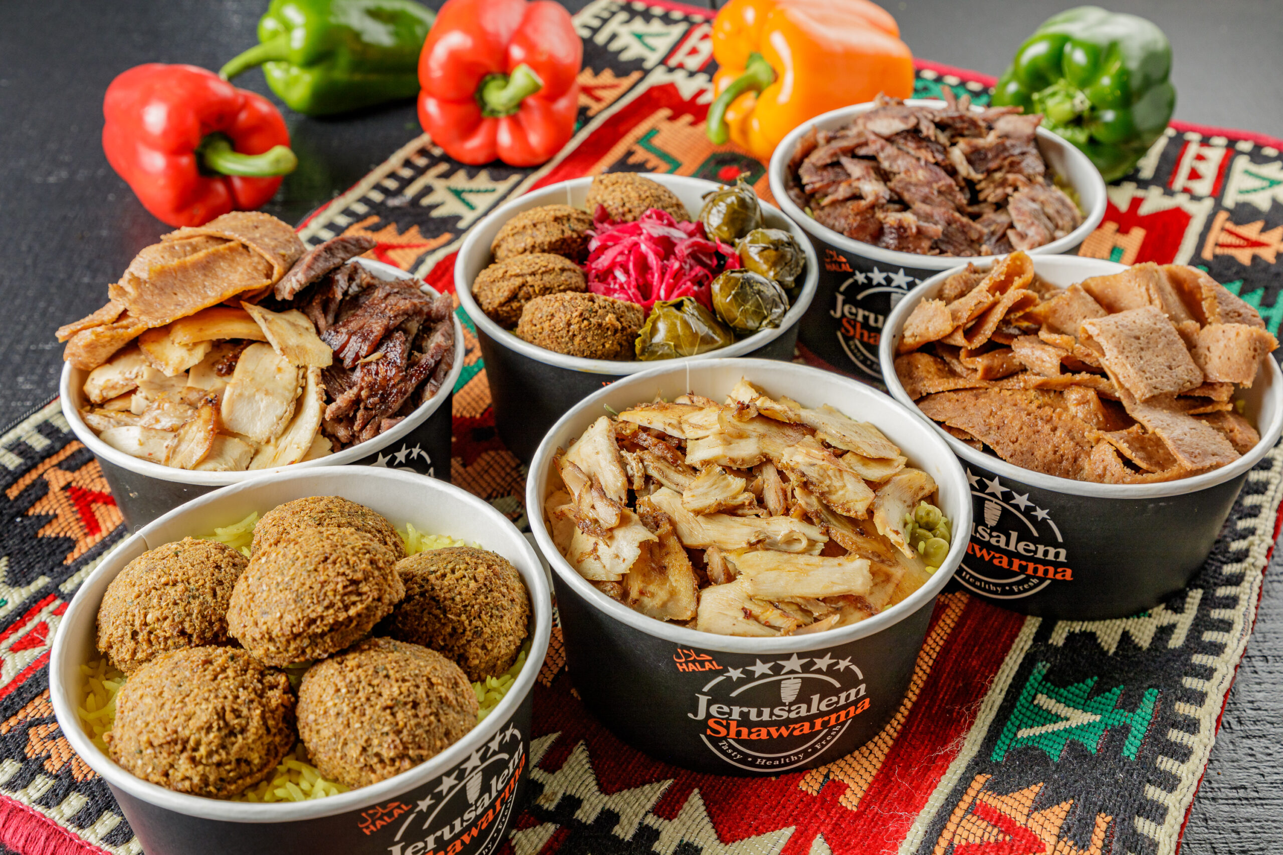 A colorful display of Middle Eastern cuisine from Jerusalem Shawarma, featuring six black branded bowls filled with a variety of dishes including falafel over yellow rice, chicken shawarma, beef shawarma, donair meat, a mix bowl of meats, and a bowl with falafel, pickled cabbage, and grape leaves. The bowls are arranged on a traditional patterned Middle Eastern cloth, with fresh red, green, and orange bell peppers in the background for decoration.
